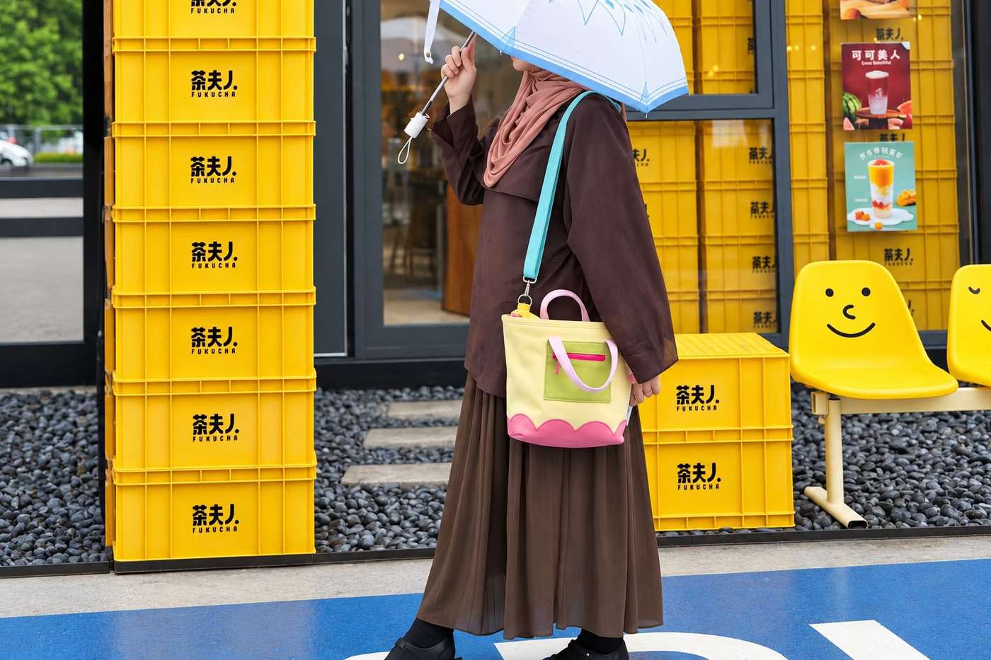 A person walking outdoors under a light blue and white umbrella, wearing a brown outfit and carrying a pink and yellow bag, with yellow crates and a city background.